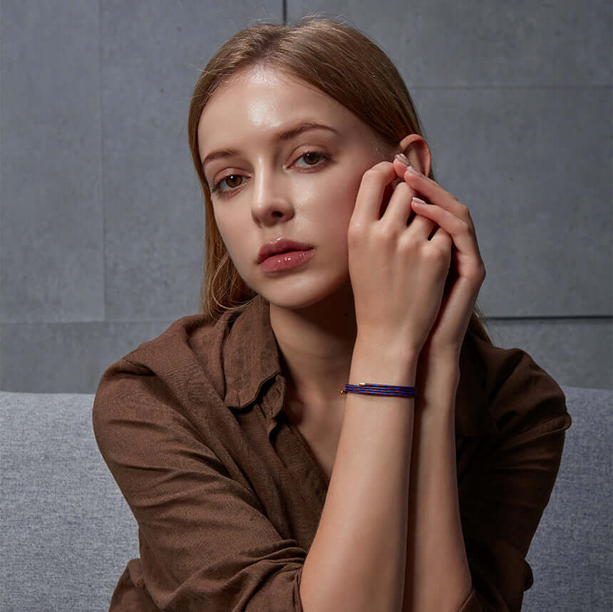 Woman wearing brown top and a blue bracelet, sitting on a gray couch, with a thoughtful expression, against a gray wall background.